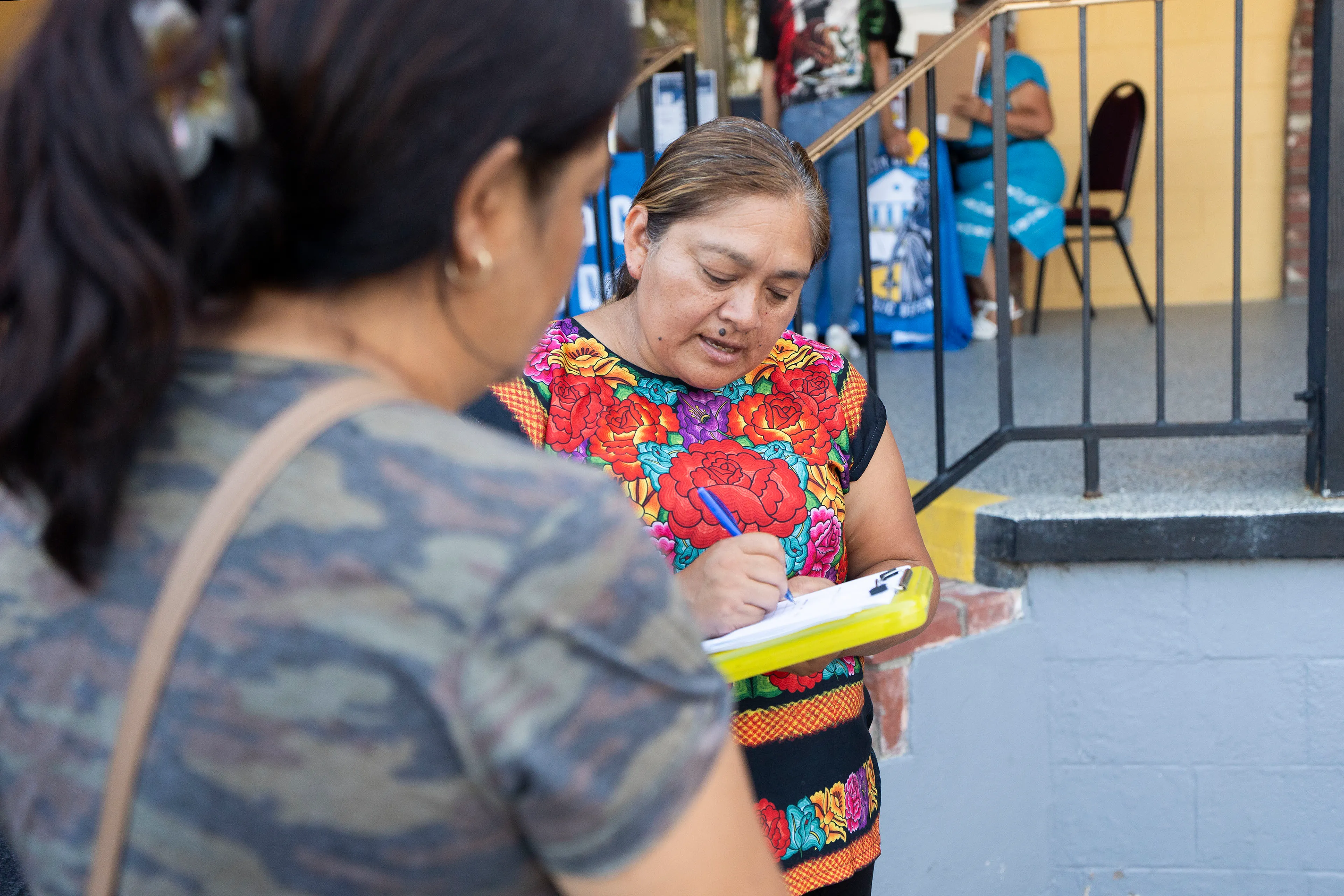 A woman writes on a clipboard as she speaks to another person whose back is to the camera.