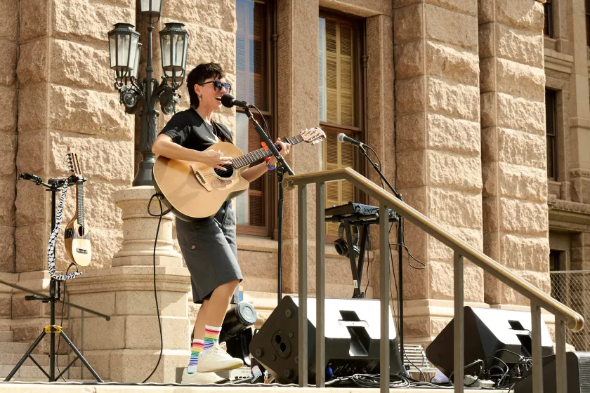 A person in a black T-shirt plays a guitar on the steps of a state building on a sunny day.