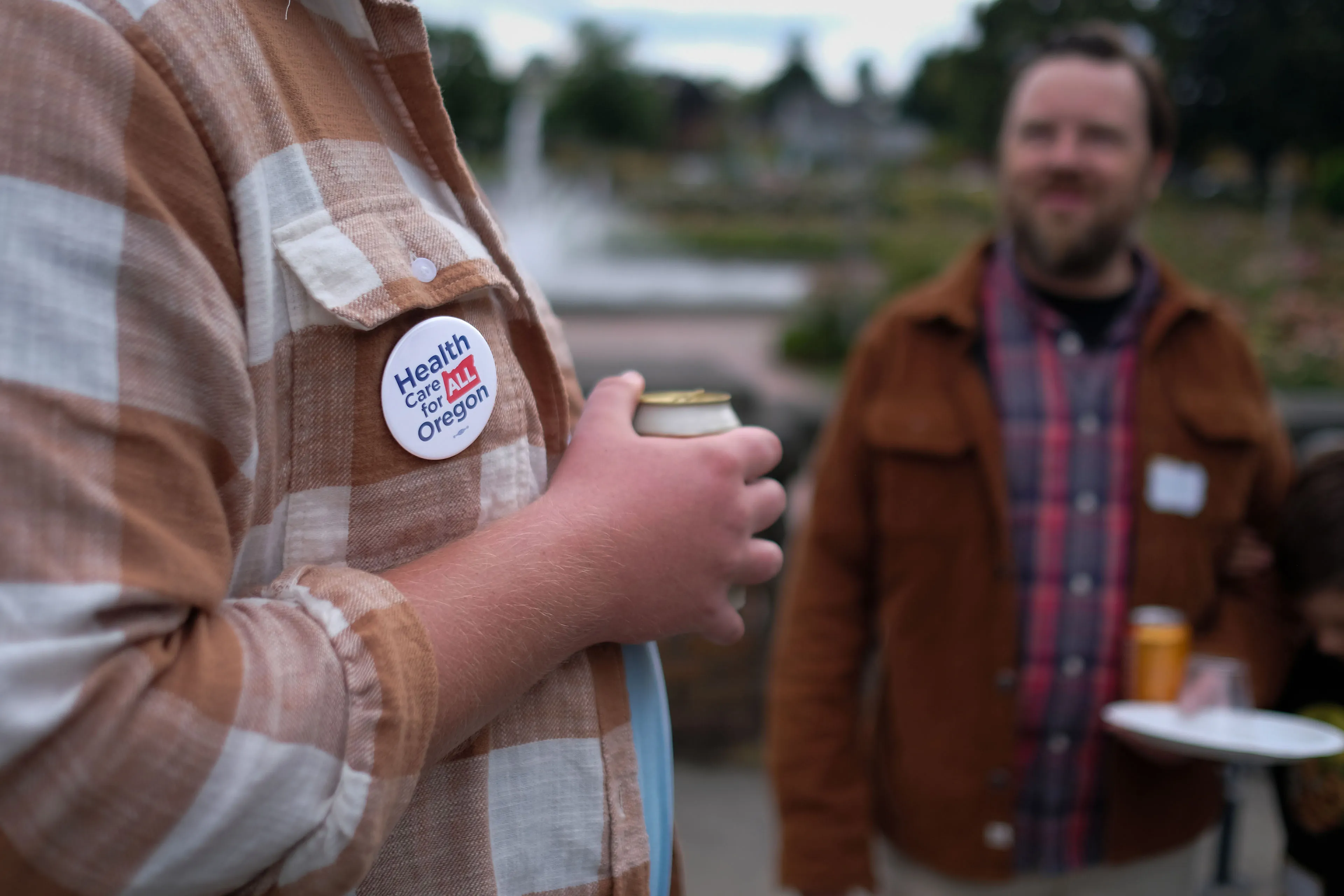 A man wearing plaid is holding a canned beverage while speaking to another man close beside him. A button is visible on the man in plaid's shirt, it says "Health Care for All Oregon."