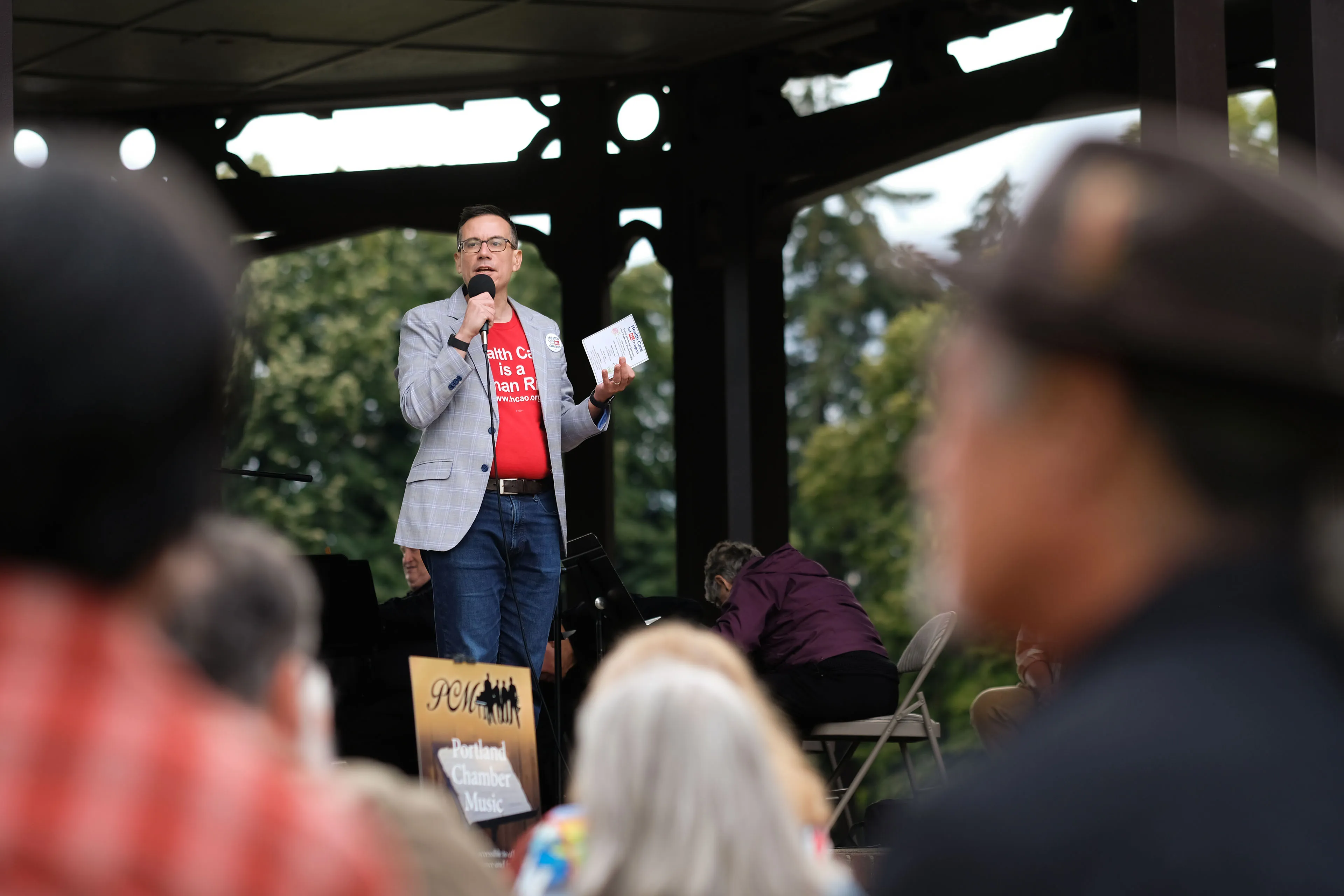 A man wearing a blazer over a bright red shirt stands in front of a crowd while speaking into a microphone in an outdoor setting.